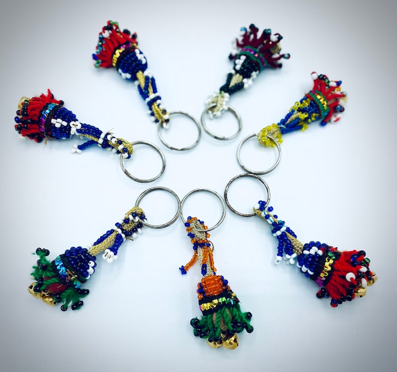 May include: Seven colorful beaded keychains with tassels hanging from silver key rings. The keychains are made of blue, red, green, yellow, and white beads.