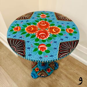 May include: A hand-painted wooden side table with a blue base and a round top. The top is painted with a floral design featuring red, orange, and yellow roses on a blue background. The base is also painted with a floral design.
