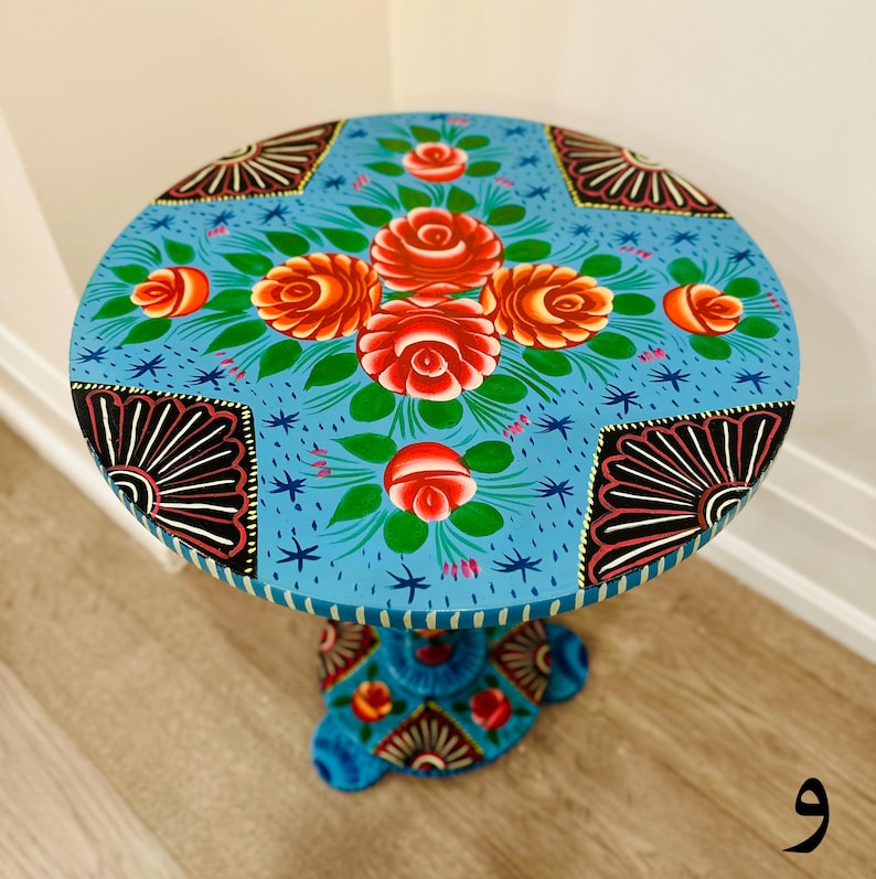 May include: A hand-painted wooden side table with a blue base and a round top. The top is painted with a floral design featuring red, orange, and yellow roses on a blue background. The base is also painted with a floral design.