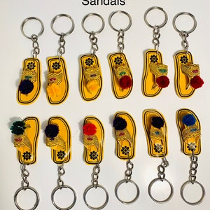 May include: Ten yellow sandal keychains with colorful pom-poms. The sandals are made of wood and have a gold-colored design. The keychains are attached to silver key rings.