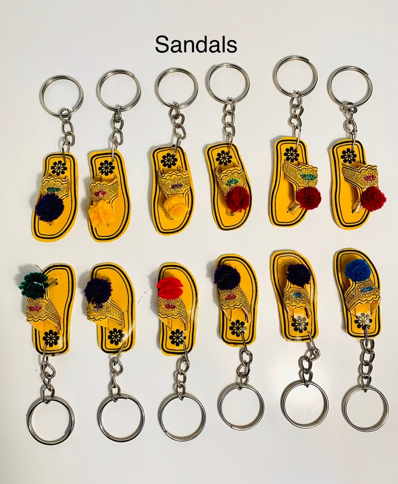 May include: Ten yellow sandal keychains with colorful pom-poms. The sandals are made of wood and have a gold-colored design. The keychains are attached to silver key rings.