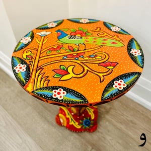 May include: A round orange table with a hand-painted design of a bird on a tree branch with colorful flowers. The table has a black base with a similar hand-painted design.
