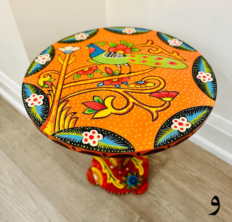 May include: A round orange table with a hand-painted design of a bird on a tree branch with colorful flowers. The table has a black base with a similar hand-painted design.