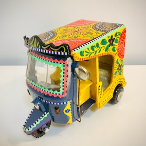 Puede incluir: Un auto-rickshaw de juguete colorido pintado a mano con un cuerpo amarillo, un frente azul y diseños florales intrincados. El rickshaw tiene un parabrisas de plástico transparente y un techo negro.
