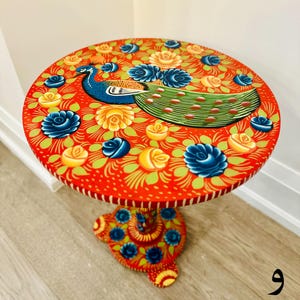 May include: A hand-painted wooden side table with a red base and a round top. The top features a peacock design with blue, green, and yellow accents. The peacock is surrounded by yellow and blue flowers.