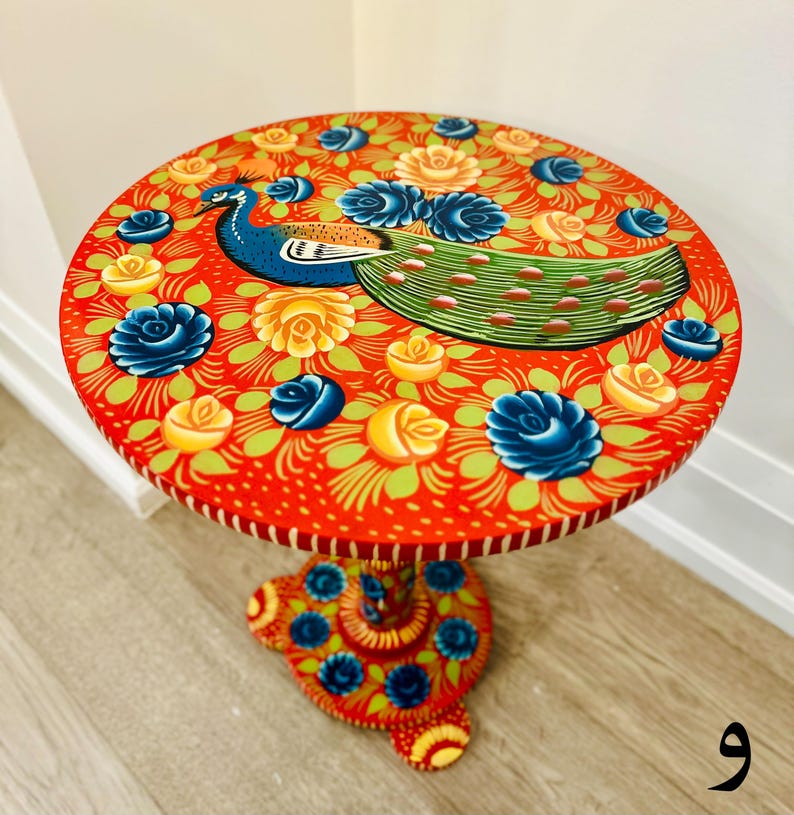 May include: A hand-painted wooden side table with a red base and a round top. The top features a peacock design with blue, green, and yellow accents. The peacock is surrounded by yellow and blue flowers.