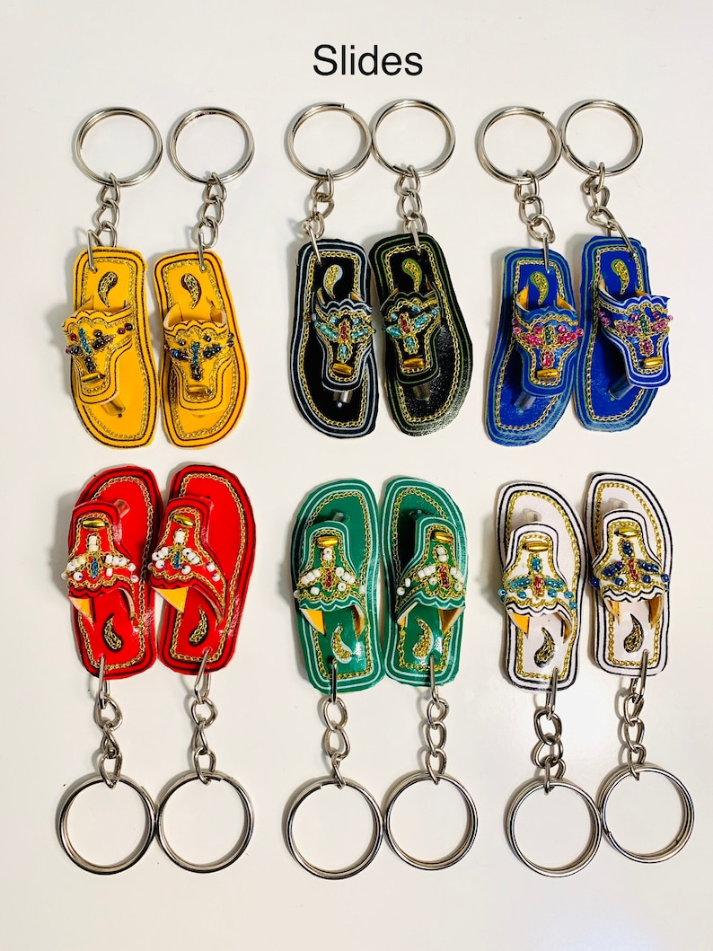 May include: Six pairs of colorful leather flip-flop keychains with intricate designs and beaded accents. The keychains are attached to silver key rings.
