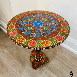 May include: A round wooden table with a hand-painted design featuring colorful flowers and leaves. The table has a brown base with a similar floral design.