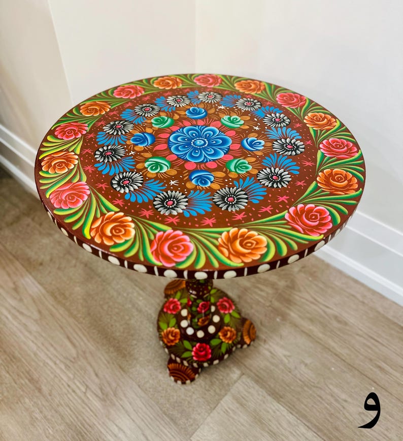 May include: A round wooden table with a hand-painted design featuring colorful flowers and leaves. The table has a brown base with a similar floral design.