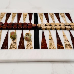 May include: A white marble backgammon board with brown and gold inlay. The board has red and brown checkers on one side and cork checkers on the other. There are two green dice with red dots on the board.