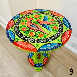 May include: A hand-painted wooden side table with a green, blue, red, and pink floral design featuring two birds. The table has a round top and a single pedestal base.