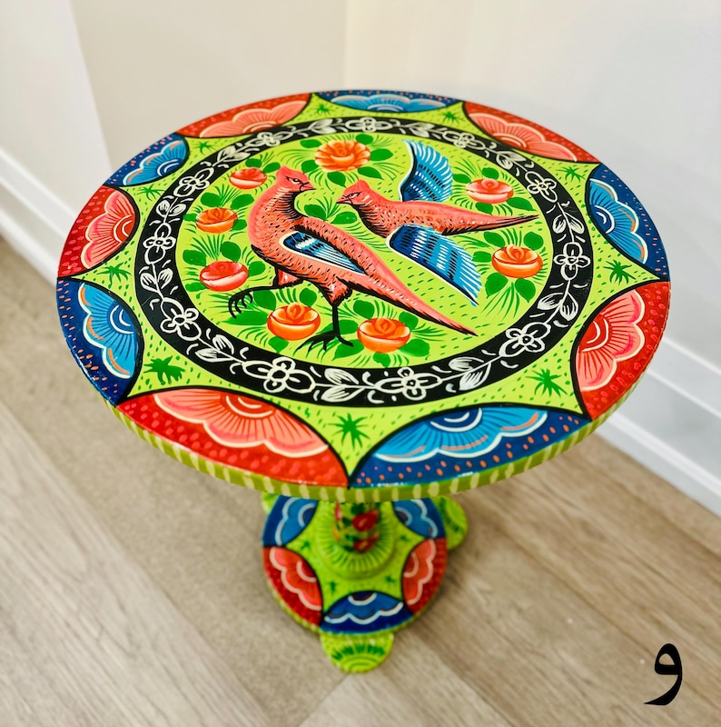 May include: A hand-painted wooden side table with a green, blue, red, and pink floral design featuring two birds. The table has a round top and a single pedestal base.