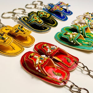 May include: A set of colorful, beaded, leather slipper keychains. Each keychain features a pair of slippers with intricate detailing and a silver key ring.