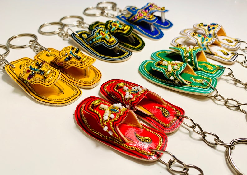 May include: A set of colorful, beaded, leather slipper keychains. Each keychain features a pair of slippers with intricate detailing and a silver key ring.
