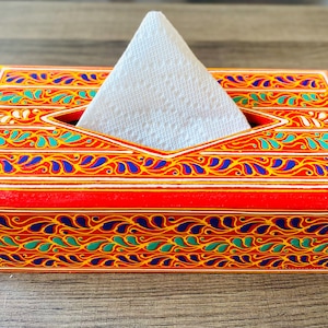 Op de afbeelding: Een rechthoekige houten tissueboxhouder, geschilderd in een levendige rode kleur met ingewikkelde handgeschilderde patronen in blauw, groen en geel. De bovenkant heeft een ruitvormige opening met een wit papieren tissue in het midden.