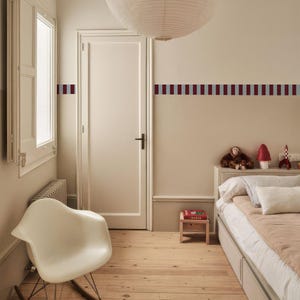 Puede incluir: Un dormitorio con una mecedora blanca, una cama con ropa de cama de colores claros y un suelo de madera. Las paredes están pintadas en un tono neutro con un patrón de rayas horizontales en colores rojo oscuro y azul claro alternados.