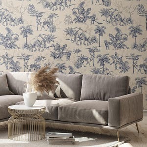May include: A beige sofa with a light gray finish, positioned in front of wallpaper featuring a repeating dark blue toile pattern of elephants, leopards, and palm trees. A gold wire side table holds a white vase with dried pampas grass.