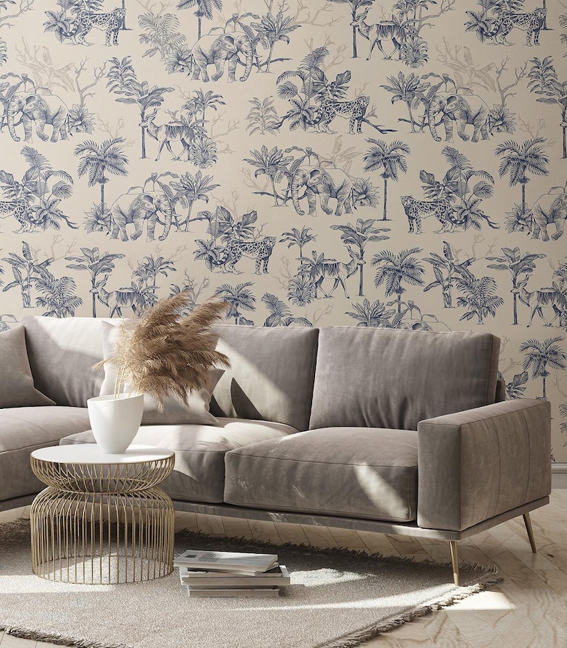 May include: A beige sofa with a light gray finish, positioned in front of wallpaper featuring a repeating dark blue toile pattern of elephants, leopards, and palm trees. A gold wire side table holds a white vase with dried pampas grass.