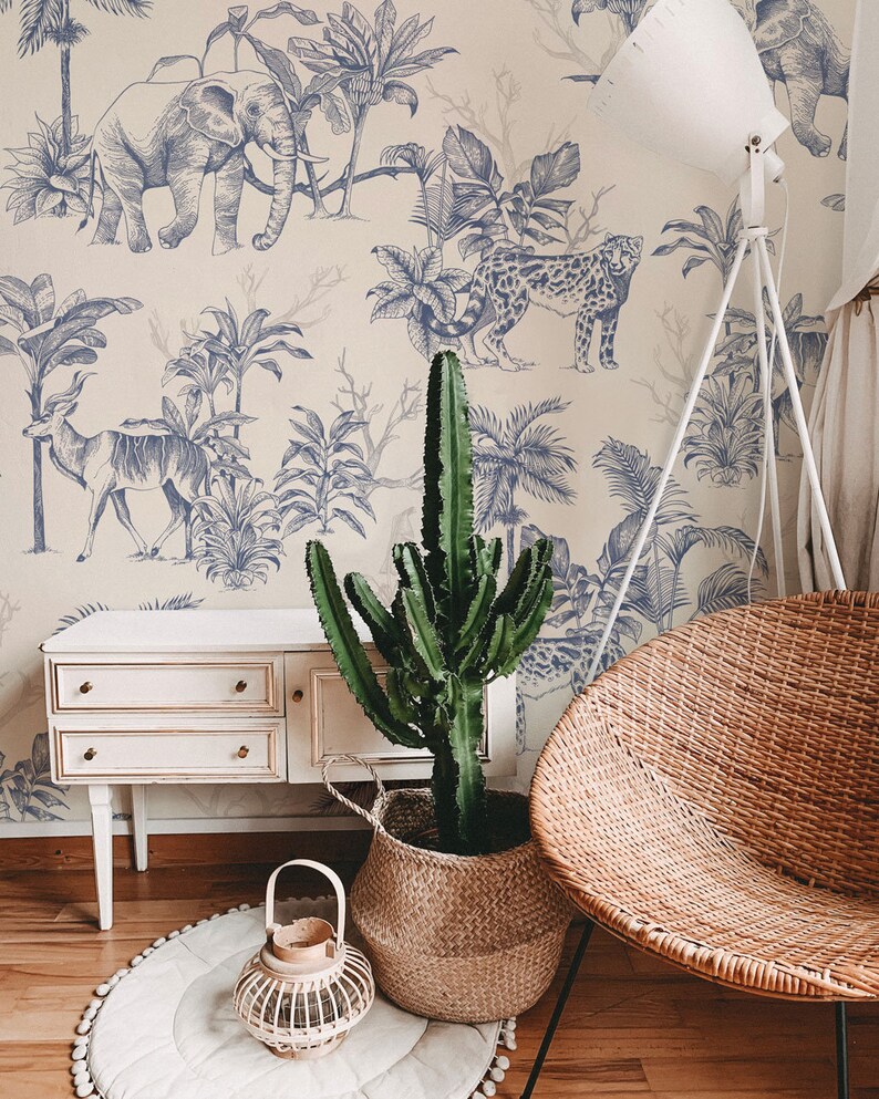 May include: A room interior featuring wallpaper with a blue ink-style jungle animal print, a white wooden cabinet, a tall green cactus in a woven basket, a round white rug, a small wooden lantern, and a round woven wicker chair with metal legs.