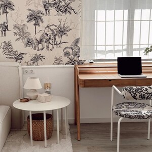 May include: A workspace featuring a wooden desk with a laptop, a white round side table with a lamp and a woven basket underneath, and a chair with a black and white patterned seat. The wall is covered in wallpaper with a black and white jungle print.