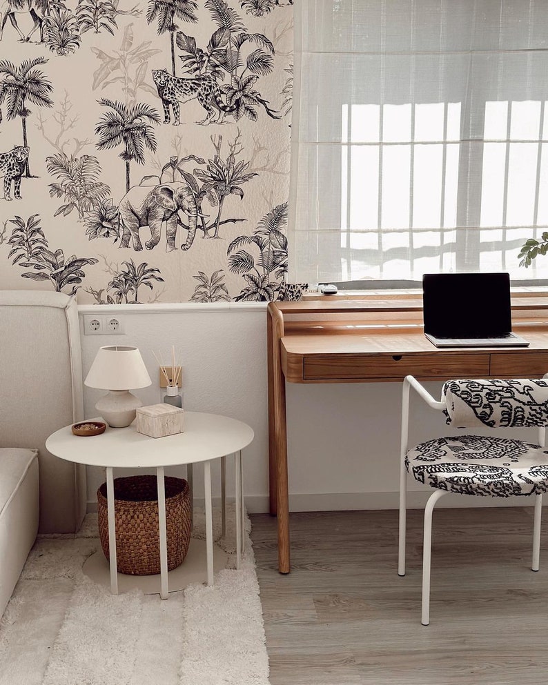 May include: A workspace featuring a wooden desk with a laptop, a white round side table with a lamp and a woven basket underneath, and a chair with a black and white patterned seat. The wall is covered in wallpaper with a black and white jungle print.
