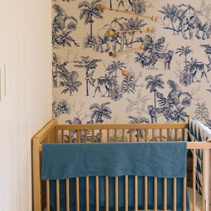 May include: A light wood crib featuring a solid blue textured blanket draped over the front rail. The wall behind the crib is covered in wallpaper with a blue and white jungle print depicting elephants, leopards, and palm trees.
