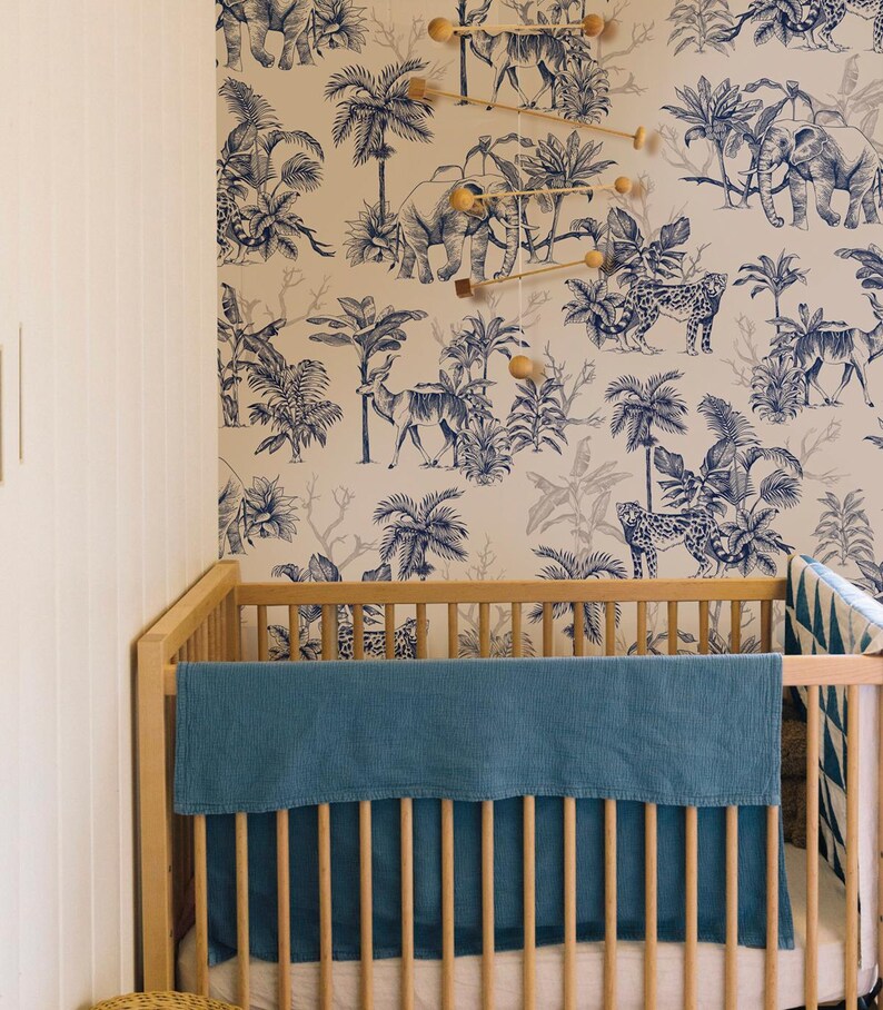 May include: A light wood crib featuring a solid blue textured blanket draped over the front rail. The wall behind the crib is covered in wallpaper with a blue and white jungle print depicting elephants, leopards, and palm trees.