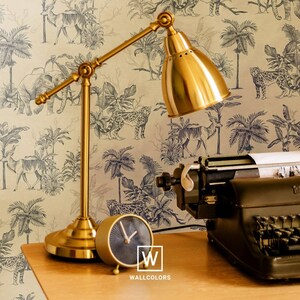 May include: A gold-toned metal desk lamp with an adjustable arm and a dome-shaped shade, a small round analog clock with a dark face, and a black vintage typewriter. The items are positioned against a wallpaper featuring a black and white jungle animal print with the text W WALLCOLORS.