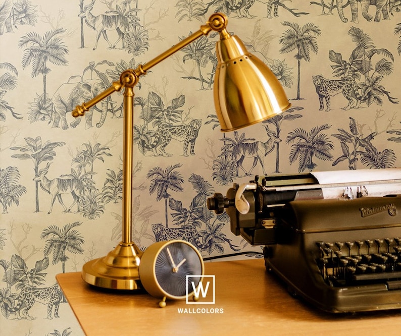 May include: A gold-toned metal desk lamp with an adjustable arm and a dome-shaped shade, a small round analog clock with a dark face, and a black vintage typewriter. The items are positioned against a wallpaper featuring a black and white jungle animal print with the text W WALLCOLORS.