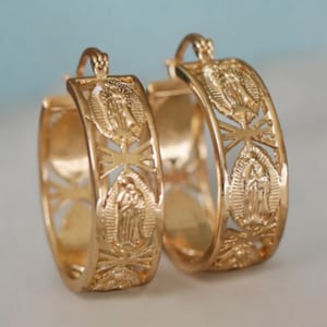 May include: A pair of gold-colored hoop earrings. Each earring features a detailed religious design with a central figure and surrounding decorative elements. The earrings have a polished finish and a secure clasp.