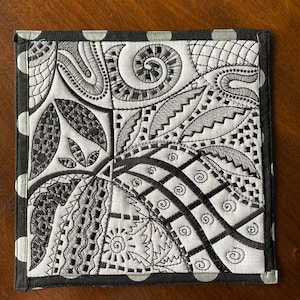 May include: Black and white embroidered fabric coaster with a geometric pattern. The coaster has a black and white polka dot border.