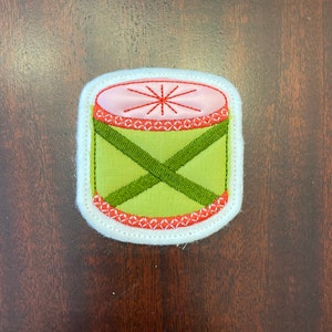 Op de afbeelding: Een groene en rode geborduurde vilt trommel patch met witte stiksels rond de randen.