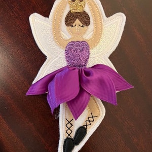 Pode incluir: Um enfeite de bailarina de feltro roxo e branco com uma coroa dourada, cabelo castanho e sapatos pretos. A bailarina está usando um vestido roxo com um laço branco.