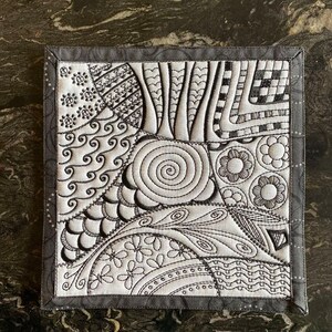 May include: A black and white quilted square featuring a range of intricate patterns, including swirls, flowers, and geometric shapes. The quilt is framed with a black border.