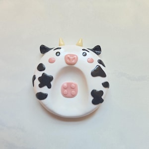May include: A round white ceramic spoon rest shaped like a cow face with black spots, small yellow horns, pink cheeks, and a pink snout. The center features a raised pink square detail with four small circles, resembling a cow udder pattern.