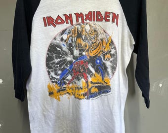 May include: White and black raglan-style t-shirt featuring the band name "Iron Maiden" in red letters. The graphic depicts a monster with flames and lightning. The sleeves are black, and the body is white.