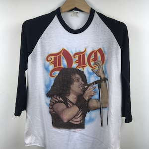 Puede incluir: Camiseta de estilo raglán blanca y negra con un gráfico de la banda Dio. La camiseta presenta un retrato del cantante y el logotipo de la banda en rojo y dorado. Las mangas son negras y la camiseta está colgada de una percha de madera.