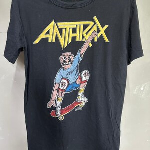 Puede incluir: Camiseta negra con el nombre de la banda "ANTHRAX" en letras amarillas sobre un skater de dibujos animados. El skater lleva pantalones cortos azules, una camisa azul y rodilleras y coderas, montando un monopatín rojo.