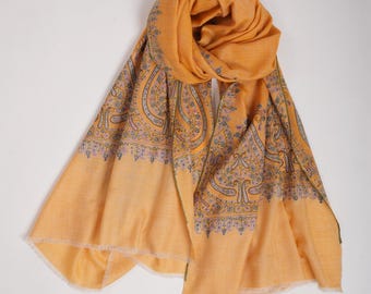 May include: A yellow paisley patterned shawl with a fringed edge. The shawl is folded and draped over a white surface.