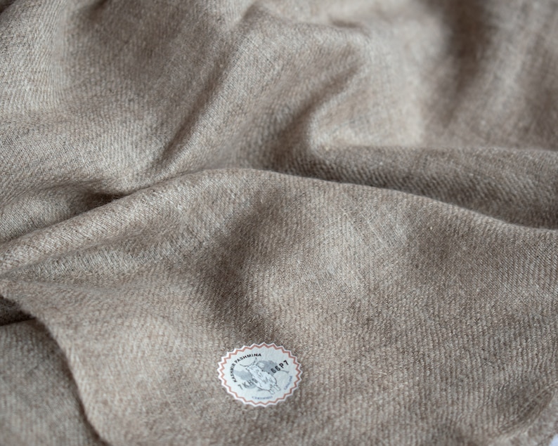 May include: A close-up of a beige wool scarf with a small round label that says "Cashmere" and has a picture of a sheep on it.