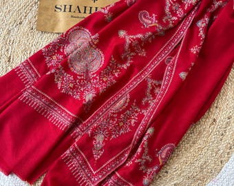 May include: A red shawl with intricate paisley and floral patterns. The shawl is made of fine fabric and has a delicate fringe. The shawl is folded and displayed on a woven rug. The text "SHAHI Handcrafted" is visible on a brown paper bag in the background.
