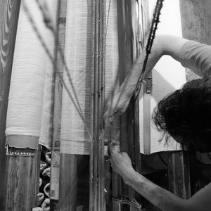 May include: A person working on a loom, weaving fabric. The loom is made of wood and has a white fabric being woven on it. The person is using their hands to guide the fabric through the loom.