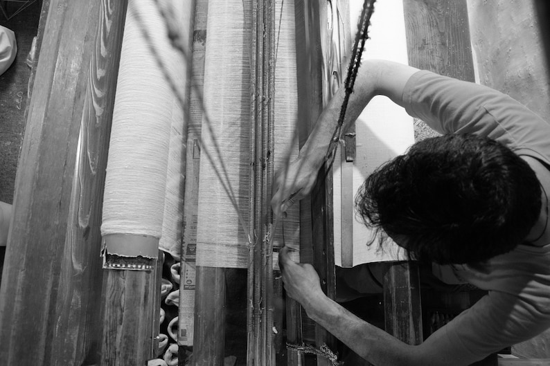 May include: A person working on a loom, weaving fabric. The loom is made of wood and has a white fabric being woven on it. The person is using their hands to guide the fabric through the loom.