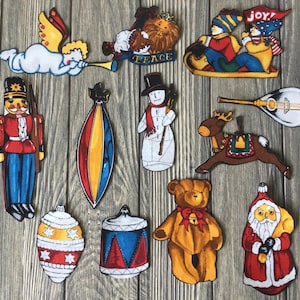 以下が含まれることがあります： 雪だるま、テディベア、ナッツクラッカー、トナカイ、楽器、ドラム、クリスマスツリーのオーナメント、サンタクロース、天使、子供2人を乗せたそりなど、さまざまなクリスマスのテーマを特徴とする、ヴィンテージのクリスマスオーナメント12個セット。オーナメントは布製で、手描きのような外観です。