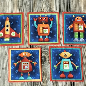 May include: Five colorful fabric squares with a blue background and a red border. Each square features a different cartoon robot or a rocket ship. The robots are orange, red, and green, with star accents. The rocket ship is red and orange.