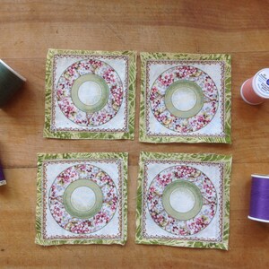 May include: Four square fabric coasters with a floral design and a green border. The coasters are arranged in two rows of two. There are four spools of thread in different colors: green, purple, pink, and blue.