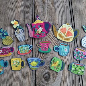 May include: A collection of colorful, hand-drawn tea-themed cutouts, including teapots, mugs, and vases with floral and bird designs. The cutouts are arranged on a wooden background, showcasing a whimsical and artistic aesthetic.