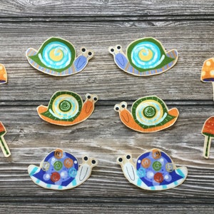 May include: Six colorful snail and mushroom cutouts on a woodgrain background. The snails are painted in shades of blue, green, orange, and yellow. The mushrooms are orange and yellow with white spots.