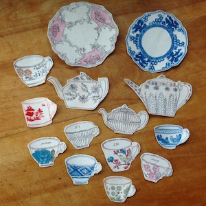 May include: Assortment of paper cutouts of teacups, teapots, and plates with floral and geometric designs. The color palette includes white, pink, blue, and red. The cutouts are arranged on a wooden surface.