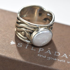 Puede incluir: Un anillo ancho de plata con una perla blanca ovalada grande. El anillo tiene un diseño texturizado en forma de ola alrededor de la banda. El anillo está sobre una caja marrón con el texto "SILPADA find yourself in it."